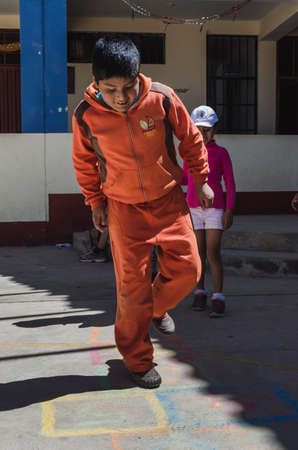 Yungay, Peru, August 8, 2014: portrait of indigenous boy with orange tracksuit jumping playing hopscotch and girl behind looking in schoolyardのeditorial素材