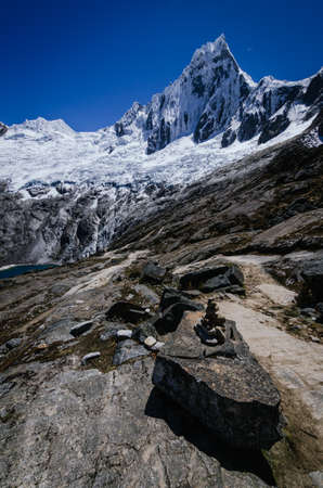 pile of stones in the foreground with high snowy mountain of Taulliraju in the background, on the trekking of the quebrada santa cruz in peruの写真素材