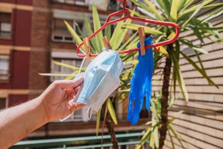 Caucasian person's hand holding surgical mask hanging next to other covid protection elements from a red hanger using tweezers on a terrace, horizontalの写真素材