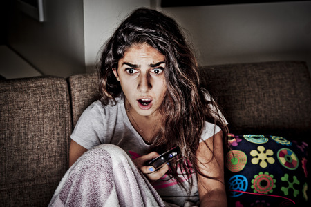 Scared young woman watching tv. Brunette girl sitting on couch with remote controlの写真素材