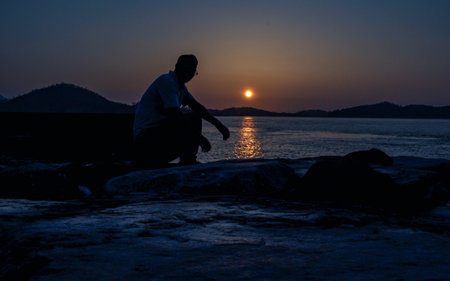 Silhouette of a man sitting on a rock by the sea at sunset