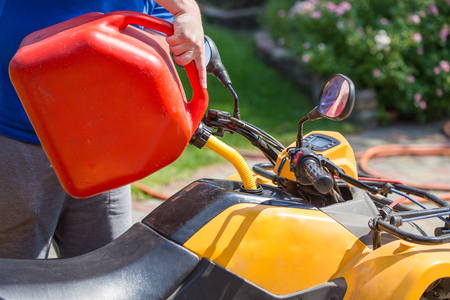 Caucasian man in sport protective goggles holds red canister and pours gasoline into ATVs quadbike tank. Refueling process. Maintenance and serviceの写真素材