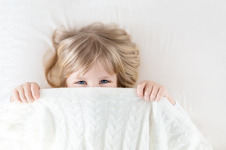 Little girl peeking out blanket on bed. Cute kid smiling and hiding under knitted cover. Palyful and mischievous eyes. Hide-and-seek. Children having fun playing active games. Happy childhood conceptの写真素材