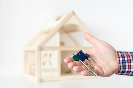 Agent's hand hold keys against house model on background. 
Realtor sale offer.  Real estate investment proposition. Buying new house concept.の写真素材