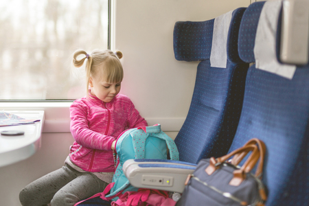 Little girl travel  by train. Kid sitting in comfortable chair and looking in backpack. Things to take with on railroad trip with children concept.の写真素材