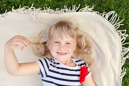 Little cute blond girl lying on blanket over green grass lawn and smiling. Adorable child having fun outdoors. Happy family, parenthood and hildhood conceptの写真素材