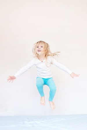 Happy kid jumping over bed. Cute little blond girl having fun indoors. Happy and careless childhood conceptの写真素材