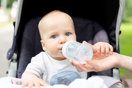 Little kid sitting in stroller and drinking water from bottle during walk at hot summer day. Mom...