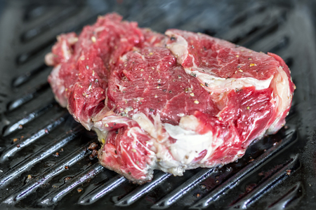 Close-up raw tasty fresh rib eye steak on grill cast iron pan. Organic beef meat seasoned with ground pepper coocked on barbequeの写真素材