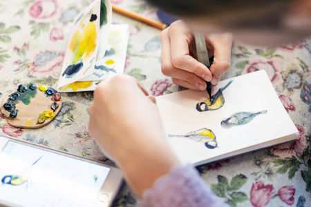 Teenager girl drawing bird in small paper drawing-pad. Close-up kid artist painting small picture on paper notepad with brush. Children creativity concept.の写真素材