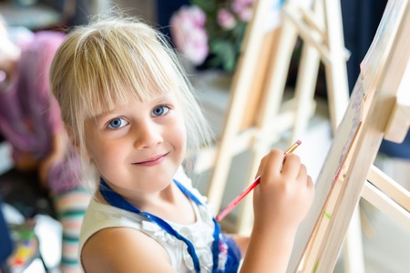 Cute blond smiling girl painting on easel in workshop lesson at art studio. Kid holding brush in hand and having fun drawing with paints. Child development concept.の写真素材