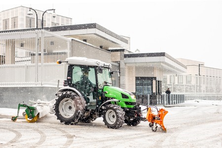 Tractor with brush and scoop cleaning snow at city street and parking during heavy snowfall at...