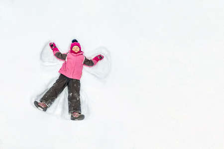Cute little kid girl in warm ski sport suit making snow angel outdoors. Kid having fun lying on snowdrift after snow storm in winter. Children outdoor activities. Winter vacation and holidays.の写真素材