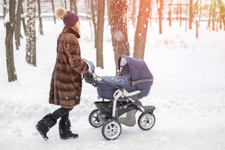 Young beautiful caucasian woman in mink fur coat walking with stroller in a forest or city park on a bright frosty winter day. Tonedの写真素材