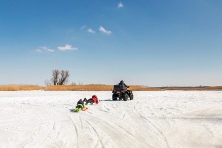 Man on ATV quadbike riding sledges with kids in tow on frozen lake surface at winter. Winter extreme sports and recreation. Children outdoor fun and activities.の写真素材