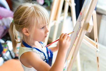 Cute blond smiling girl painting on easel in workshop lesson at art studio. Kid holding brush in hand and having fun drawing with paints. Child development concept.の写真素材