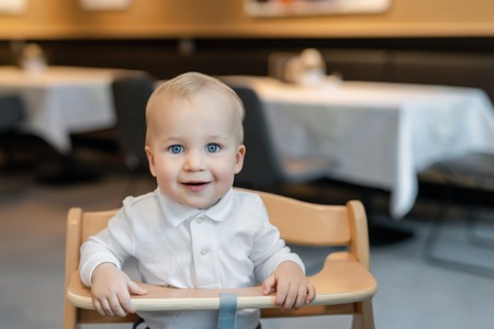 Cute little baby boy in white polo t-shirt sitting in wooden baby chair and laughing at cafe...