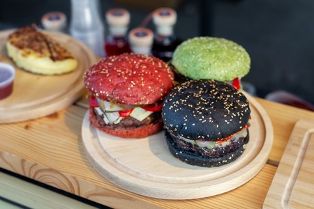 Three tasty bright multicolored beef burgers on wooden counter at food fest. amburger bun clolred with tomato, basil and cuttlefish inkの写真素材