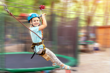 Cute little funny caucasion blond girl in helmet having fun riding rope zipline in adventure park. Children outdoor extreme sport activities conceptの写真素材