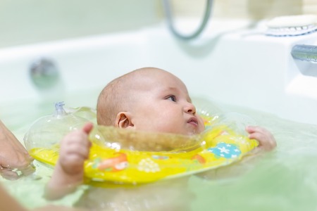 Cute little infant boy swimming with inflatable neck swim ring in bath. Baby having fun during water...