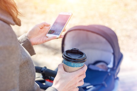 Woman holding cup of coffee to go and using smartphone during walk with baby carriage. Modern young business woman combining work and child care.の写真素材