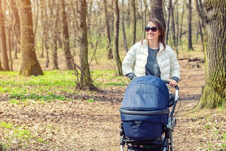 Beautiful young adult woman walking with baby in stroller through forest or park on bright sunny day. Healthy lifestyle and children healthcare. Happy childhood and parenthood.の写真素材