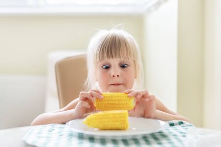 Cute blond caucasian girl holding in hand tasty boiled sweet corn cob and lokking on it with amazed...