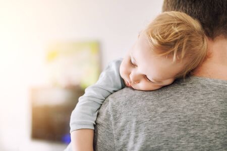 Close-up portrait of cute adorable blond caucasian toddler boy sleeping on fathers shoulder indoors. Sweet little child feeling safety daddys hand. Responsibility and childcare. happy childhood.の写真素材