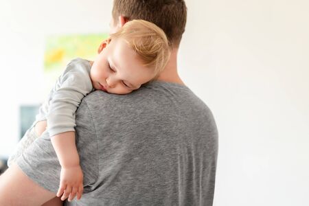 Portrait of cute adorable blond caucasian toddler boy sleeping on fathers shoulder indoors. Sweet little child feeling safety and care on daddys hand. Single father concept.の写真素材