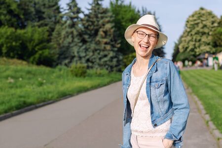 Happy young caucasian bald woman in hat and casual clothes enjoying life after surviving breast cancer. Portrait of beautiful hairless girl smiling during walk at city park after curing disease.の写真素材