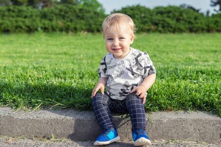 Adorable funny baby toddler boy smiling and sitting on road curb at city park with green grass on background. Cute little child having fun sitting  on pathway border.の写真素材
