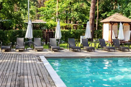 Poolside with clear blue water and loungers under umbrellas on summer day heat. Swimming pool at resort in green coniferous pine forest. Weekend destination place.の写真素材