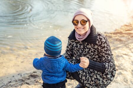 Young adult woman with cute toddler boy having funfeeding birds near lake or pond at city park on bright atumn day. Mother with adorable little son throwing bread to feed ducks near river bank.の写真素材