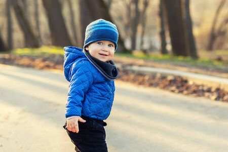 Portrait of cute funny caucasian toddler boy in blue jacket and hat enjoying walking at autumn park or forest during sunset with sun rays shining through trees on background. Baby having fun outdoors.の写真素材