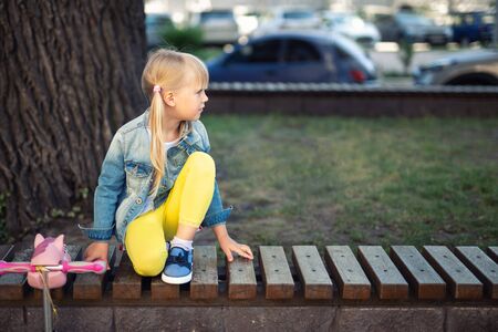 Adorable blond caucasian preschooler fashionista girl wearing jeans and bright yellow leggins sitting over wooden bench and smiling at city park or street at warm autumn day. Lifestyle child portrait.の写真素材