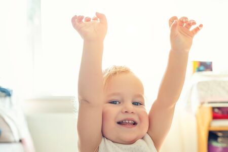 Cute adorable caucasian blond toddler boy having fun, laughing and rising hands up indoors. Cheerful child playing with positive expression at home. Happy childhood concept.の写真素材