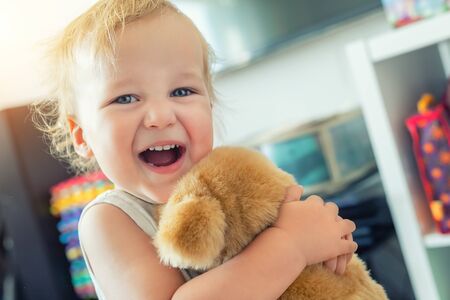 Cute adorable caucasian blond toddler boy having fun, laughing and hugging soft puppy toy indoors. Cheerful child playing with teddy bear at home. Kid friend animal toy. Happy childhood concept.の写真素材