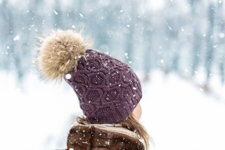 Close-up back view of young adult woman head wearing warm stylish woolen knitted handmade hat with big fluffy natural fur pompon. walking at city park or forest durin snowfall at cold winter day.の写真素材