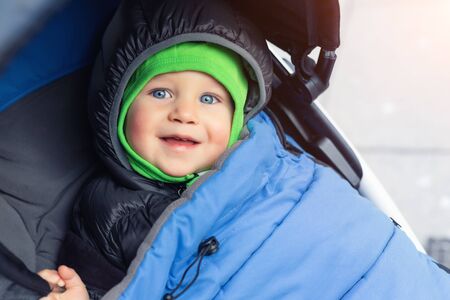 Portrait of cute caucasian toddler boy in black hooded warm insulated dawn overall jacket and blue blanket sitting in stroller and enjoy walking putdoors at cold winter day. Adorable happy childの写真素材