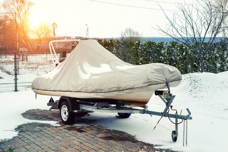 Inflatable luxury fishing motorboat wrapped in cover standing over trailer for winter period seasonal storage at backyard. Shrink-wrapped vessel winterized on parking.の写真素材