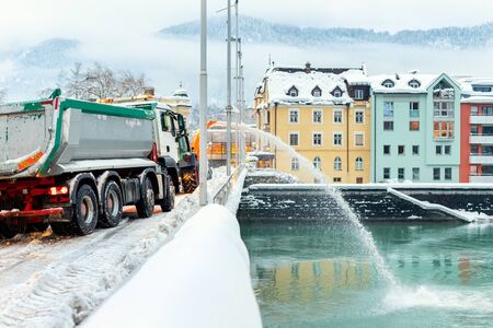 Heavy municipal services machinery removing snow from city streets. Big tractor snowblower blowing snow from bridge road into river. Cleaning streets and snow removal after snowfall at winter season.の写真素材