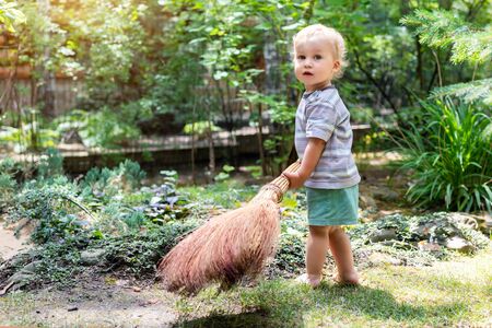 Cute adorable caucasian toddler boy playing holding broom at backyard in garden outdoors. Child little helper in t-short and shorts having fun sweeping and cleaning yard near house at countrysideの写真素材