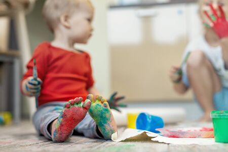 Adorable cute caucasian little blond siblings children enjoy having fun painting with brush and palm at home indoors . Cheerful happy kids smiling drawing masterpiece art picture. Messy dirty roomの写真素材