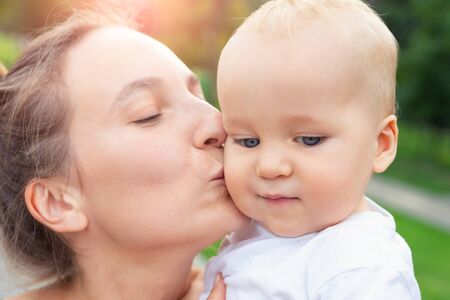 Young adult attractive beautiful caucasian mom kissing little blond toddler boy son during walk in...