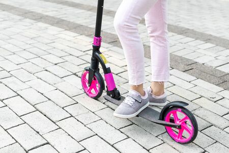 Bottom part of young adult female person wearing pink casual jeans and sneakers riding push kick...