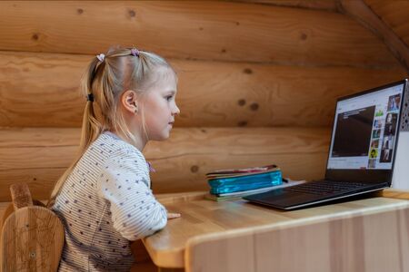 Cute adorable caucasian little blond girl siiting at desk with laptopn during online video chat scholl lesson session with teacher and class. Remote education concept. Self-isloation at quarantineの写真素材