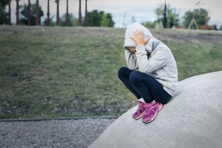 Upset sad depressed little school girl sitting alone at city street park clutching head. Upset...