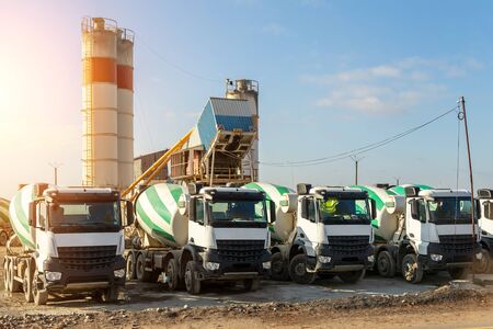 Row of many modern big mixer trucks parked against mobile temporary concrete plant factory at new asphalt road construction site morning day. Heavy machniery and industrial facilities background.の写真素材