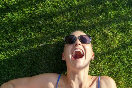 Portrait young adult bald shaved beautiful caucasian woman enjoy having fun lying on green grass lawn and smiling, Cheerful hipster feminine girl in sunglasses laughing in park or garden outdoorsの写真素材