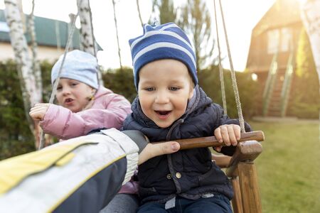 Two cute adorable playful caucasian siblings boy girl child enjoy having fun swinging wooden swing at backyard together with father. Little toddler brother and sister laughing at playing outdoorsの写真素材